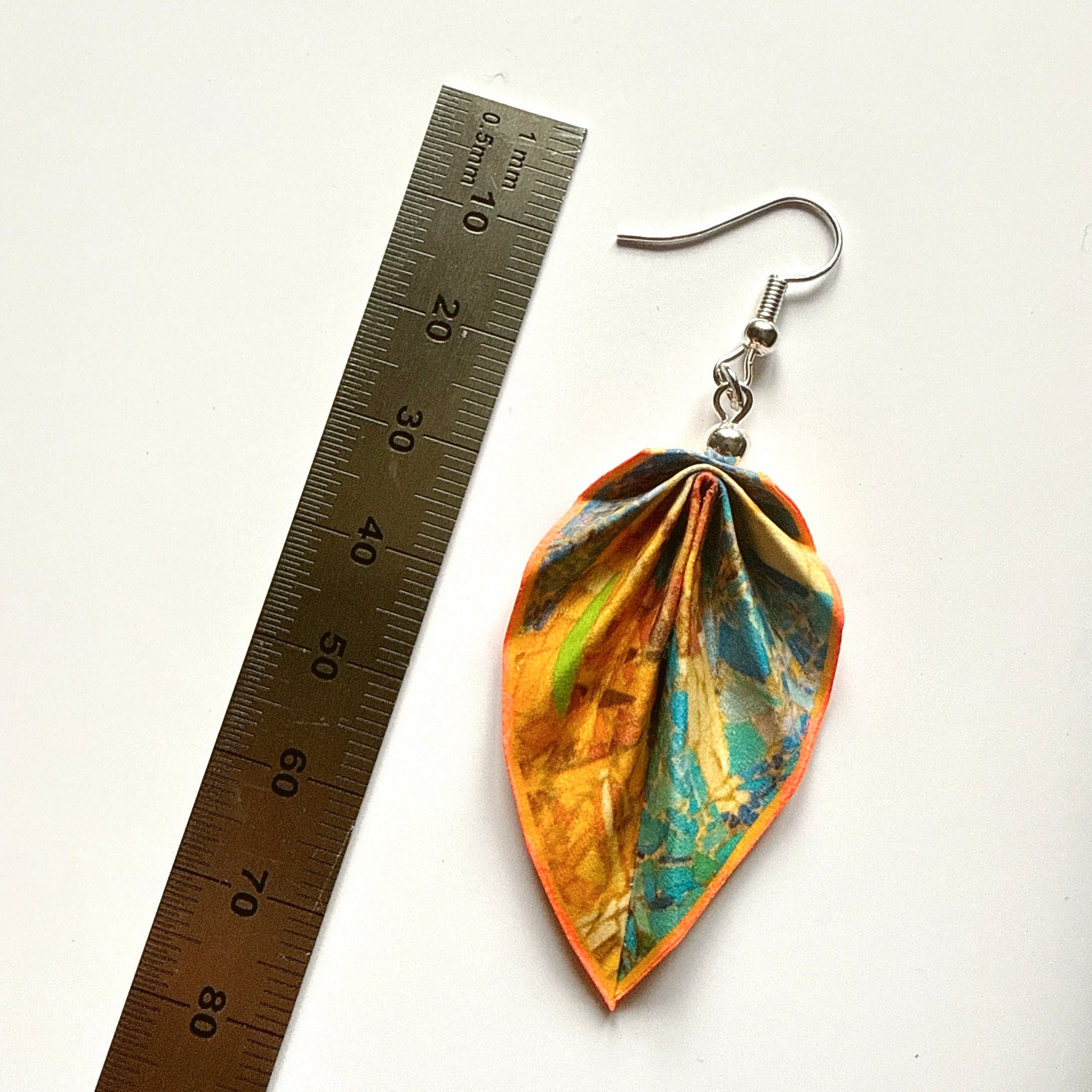 Leaf-shaped earrings with a colorful design next to a ruler on a white background