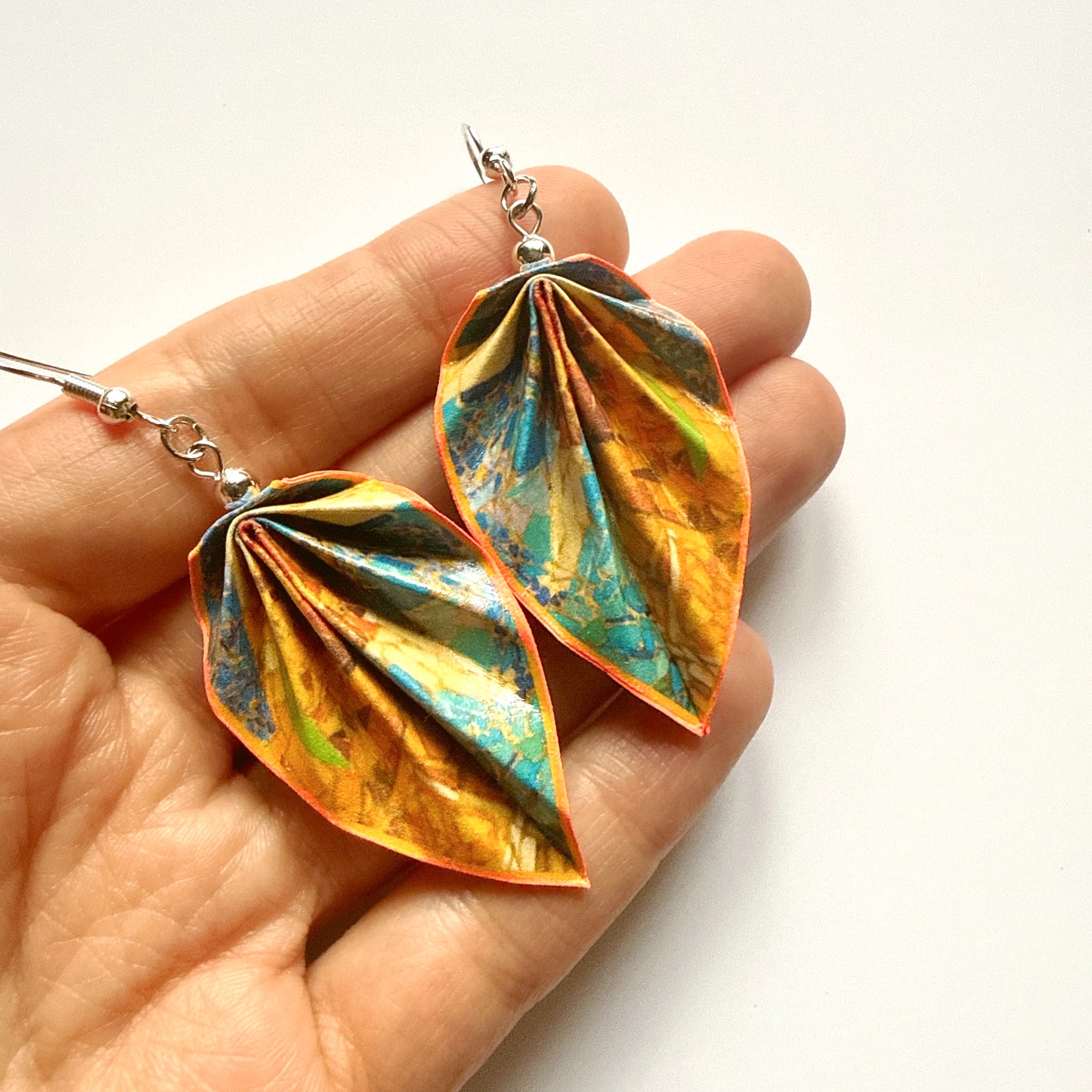 Hand holding a pair of colorful leaf-shaped earrings with a white background