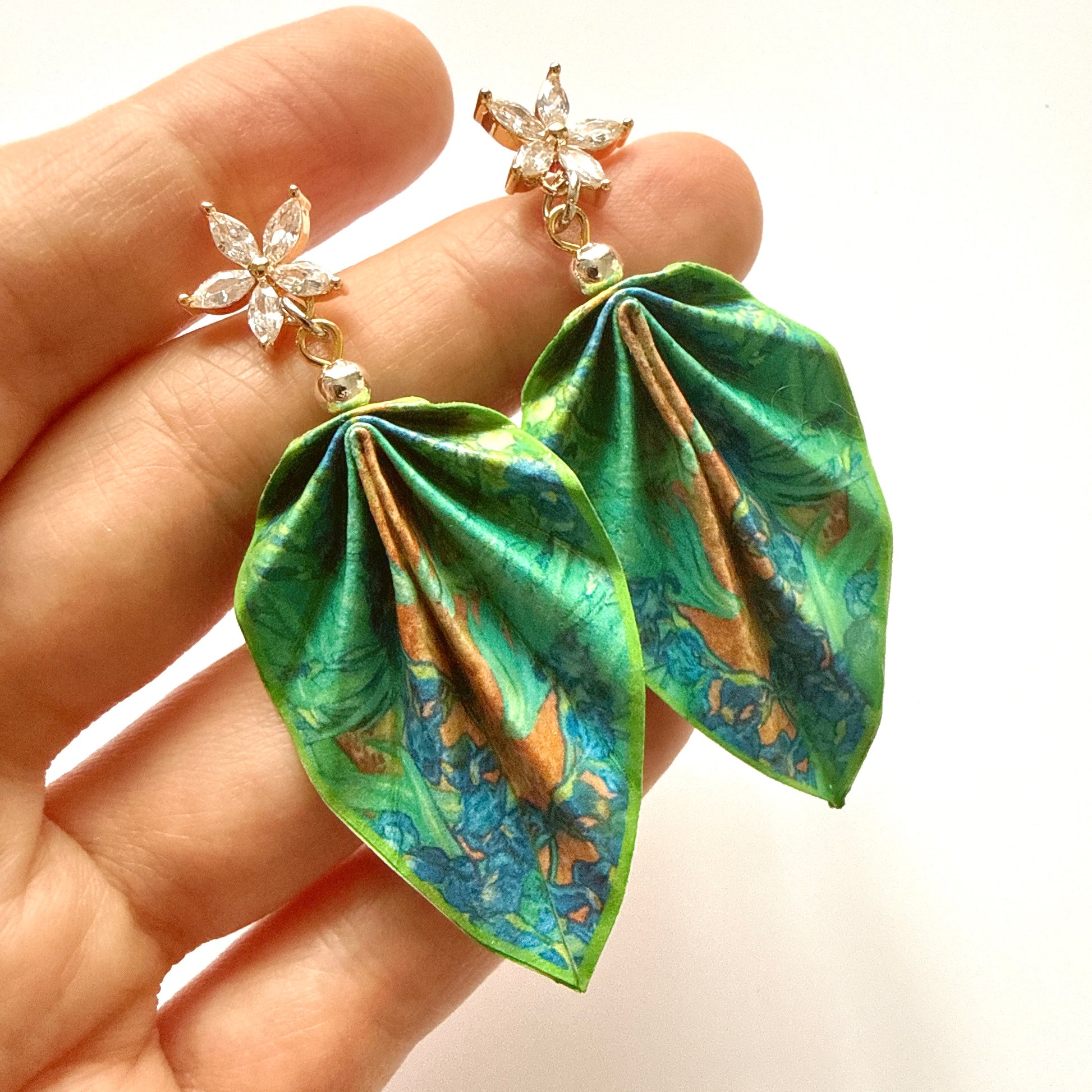 Green leaf-shaped earrings with floral details held in a hand against a white background