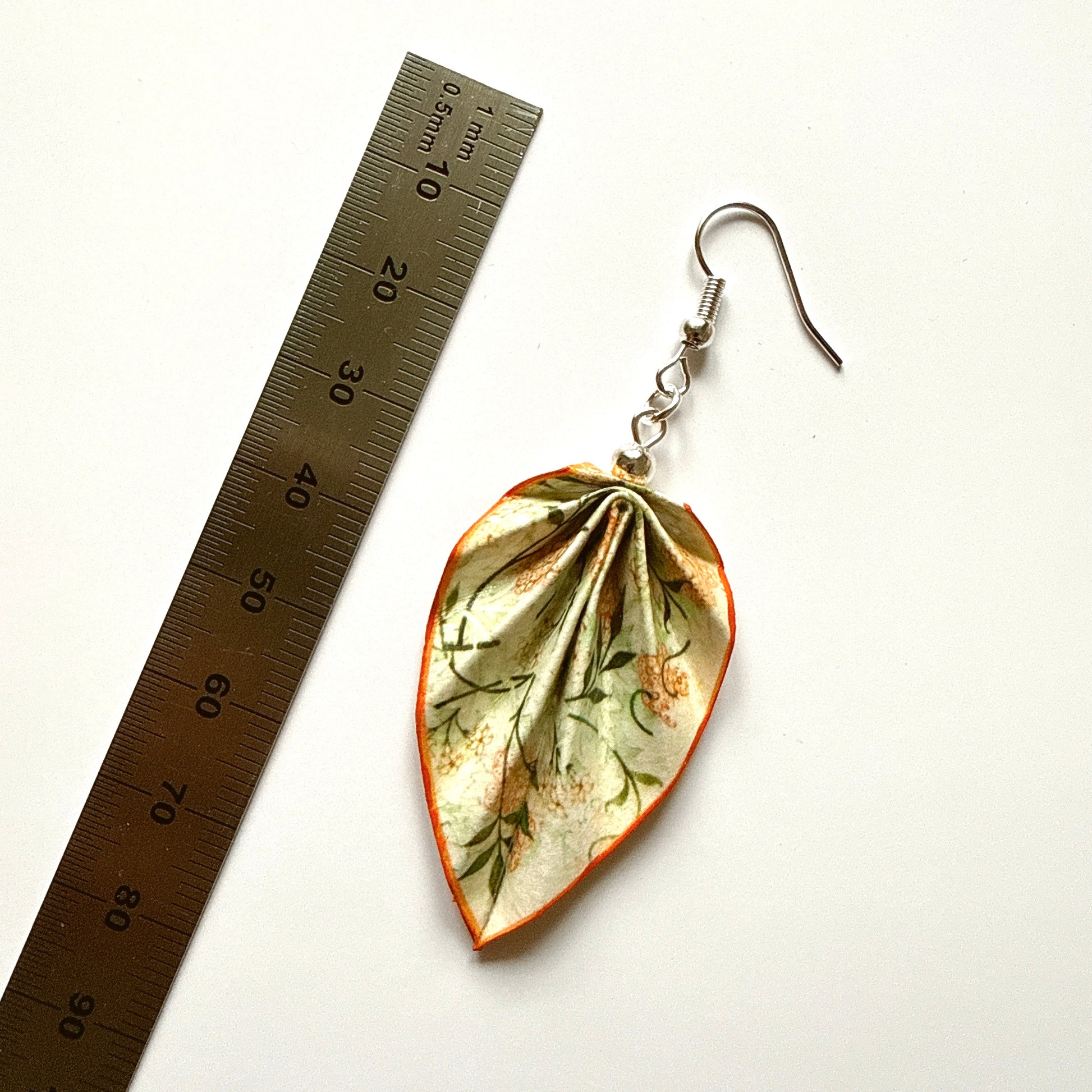 Leaf-shaped earrings with floral pattern next to a ruler on a white background