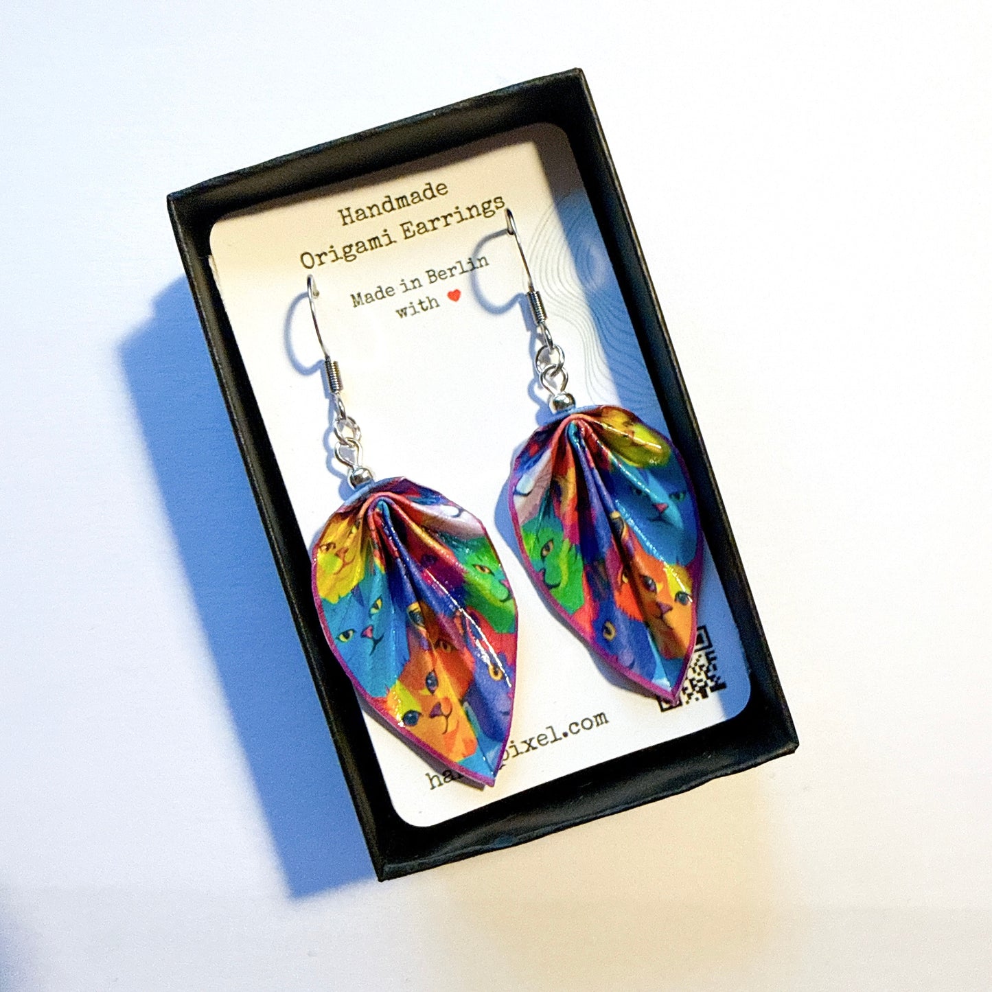 Colorful origami earrings in a black box with a card indicating 'Handmade Origami Earrings Made in Berlin'.