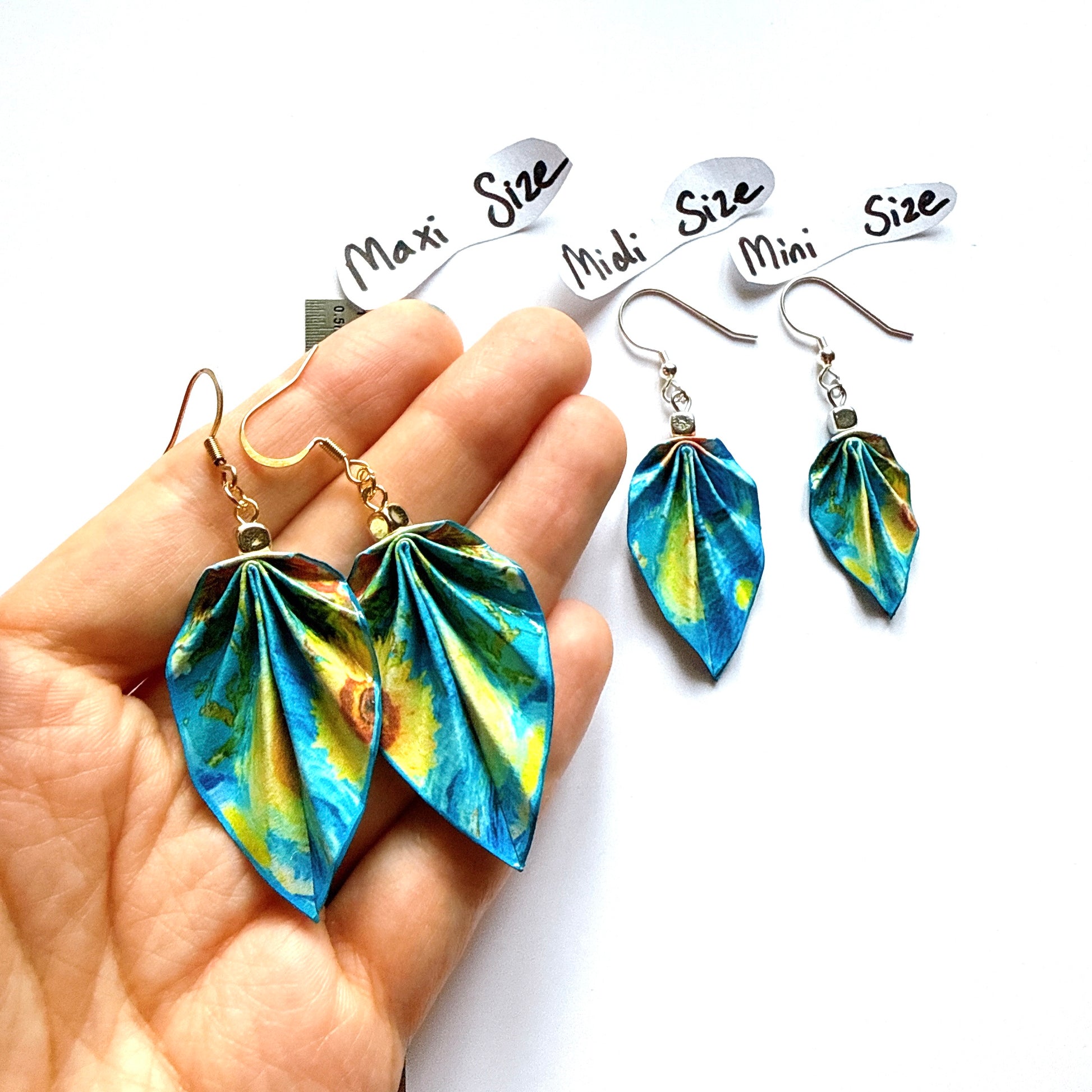 Colorful leaf-shaped earrings held in a hand with size labels in the background.