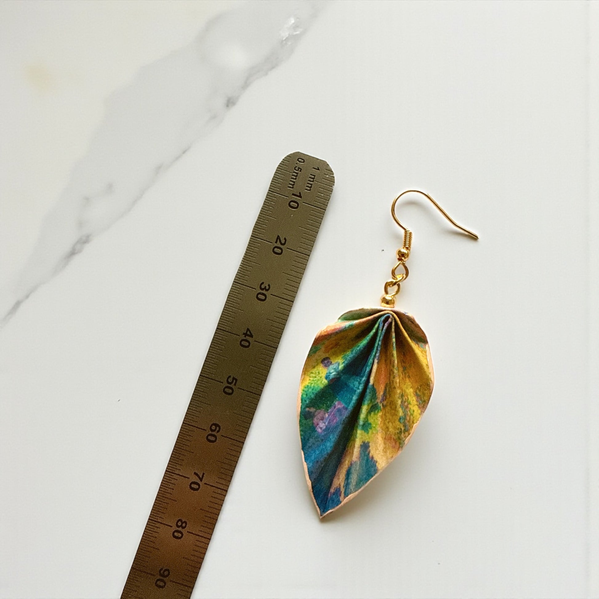 Leaf-shaped earring with gold hook next to a ruler on a marble surface